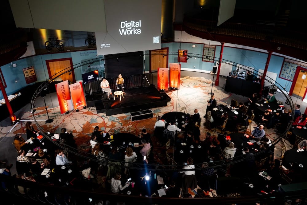 A photo taken from the balcony at Leeds City Museum looking down into the circular auditorium where the conference took place, the audience is in darkness and two people (me and Kati Price) are sitting on the stage