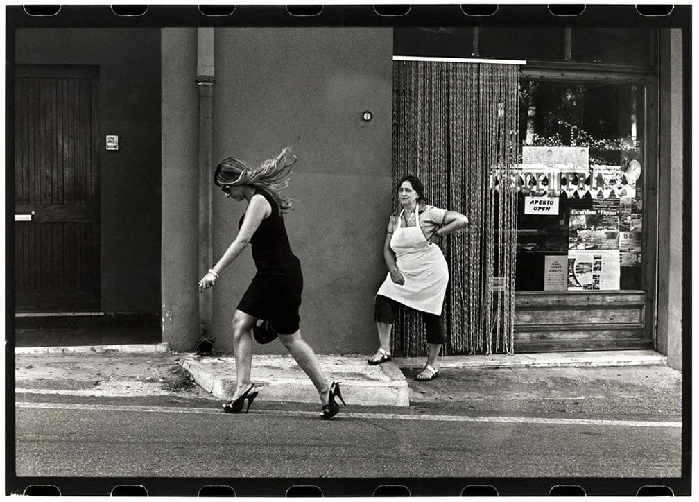 Una donna con un vestito elegante e tacchi a spillo cammina per strada, osservata dalla commessa di un negozio con un grembiule bianco.