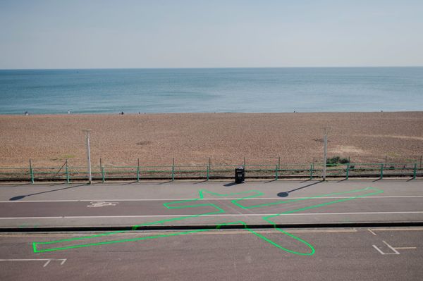 La sagome di un drone dipinta su un marciapiede di fronte a una spiaggia. Drone Shadow, un'opera di Jamse Bridle.