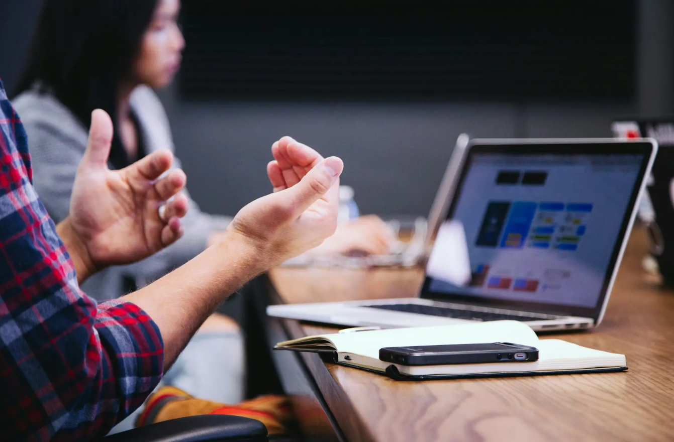 Person gesturing during a discussion with laptop and notebook on table.