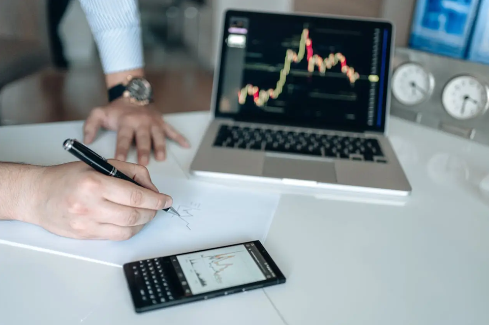 Person analyzing financial chart on paper with laptop and calculator showing stock graphs.