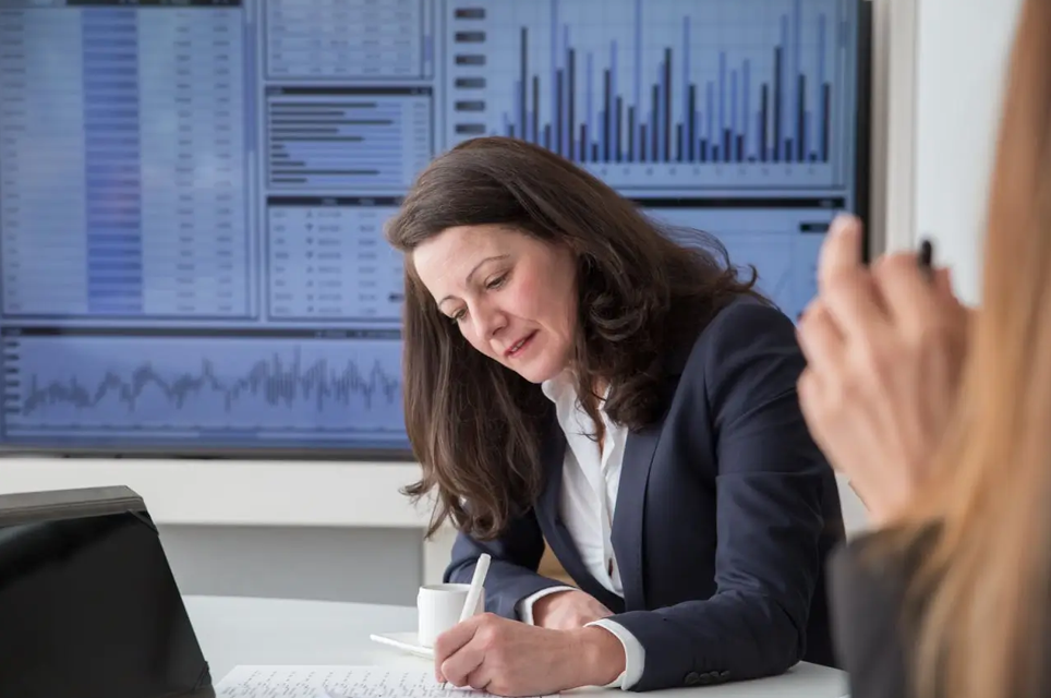 Business professional reviewing documents in a meeting room with financial charts and analytics displayed on a large screen in the background.