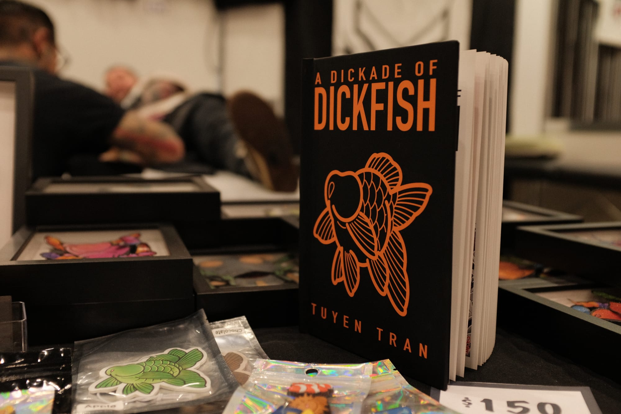 A black book titled A Dickade of Dickfish by Tuyen Tran, with an orange goldfish on the cover, stands on a table beside fish stickers and a 150 dollar price card.