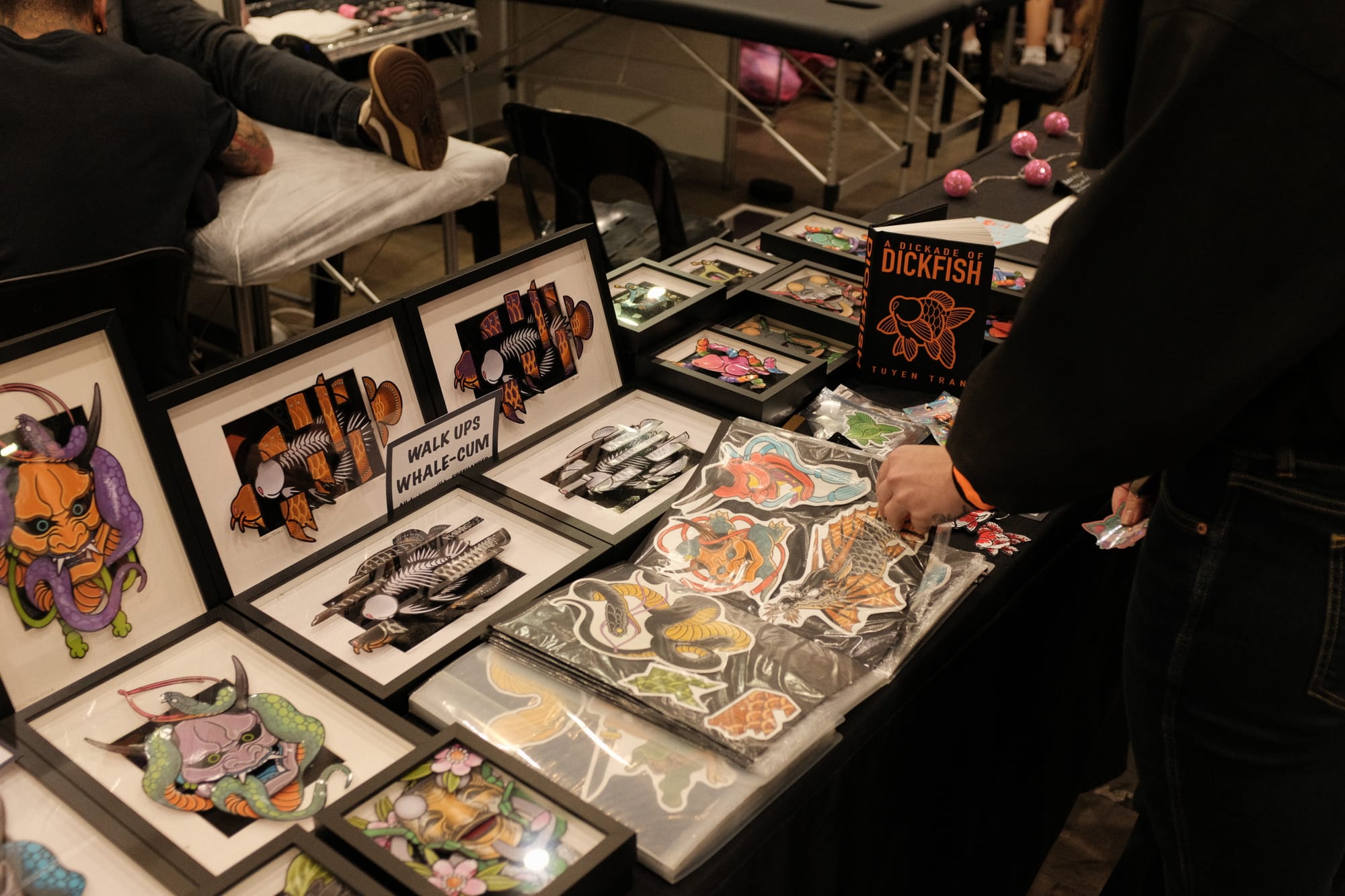 A tattoo convention booth table covered with framed Japanese-style flash prints, stickers and a book titled A Dickade of Dickfish by Tuyen Tran.