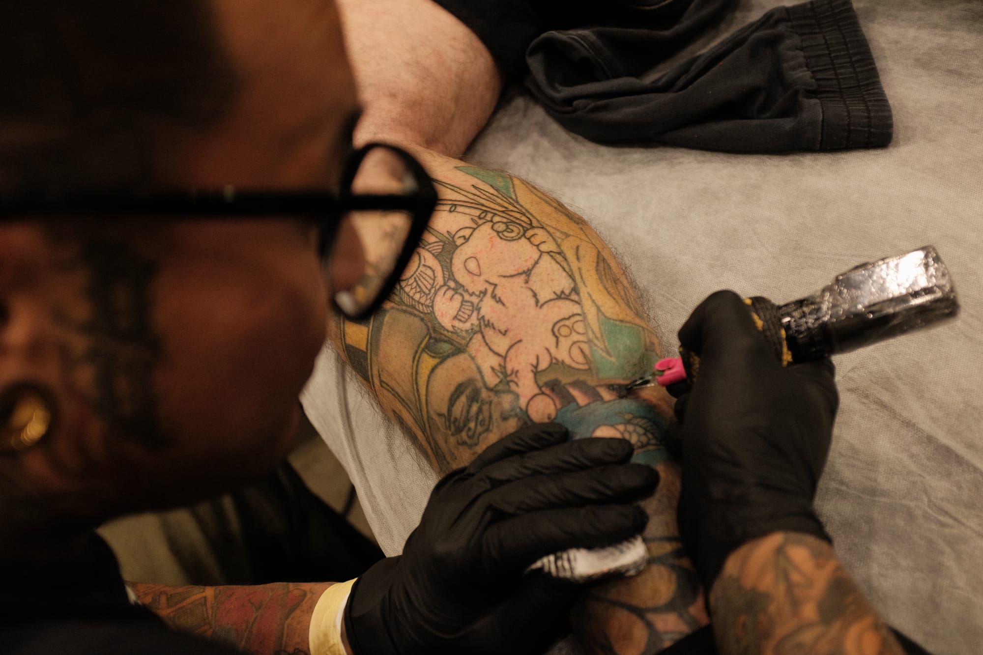 Overhead view of a bespectacled tattoo artist working a colour cartoon piece on a client's thigh with a tattoo machine, both arms heavily tattooed.