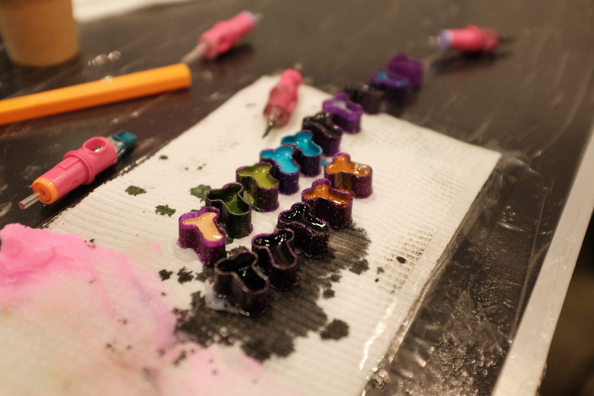 A row of small tattoo ink caps filled with green, blue, orange, black and flesh-tone ink sits on a paper towel beside pink tattoo machine grips on a workstation.
