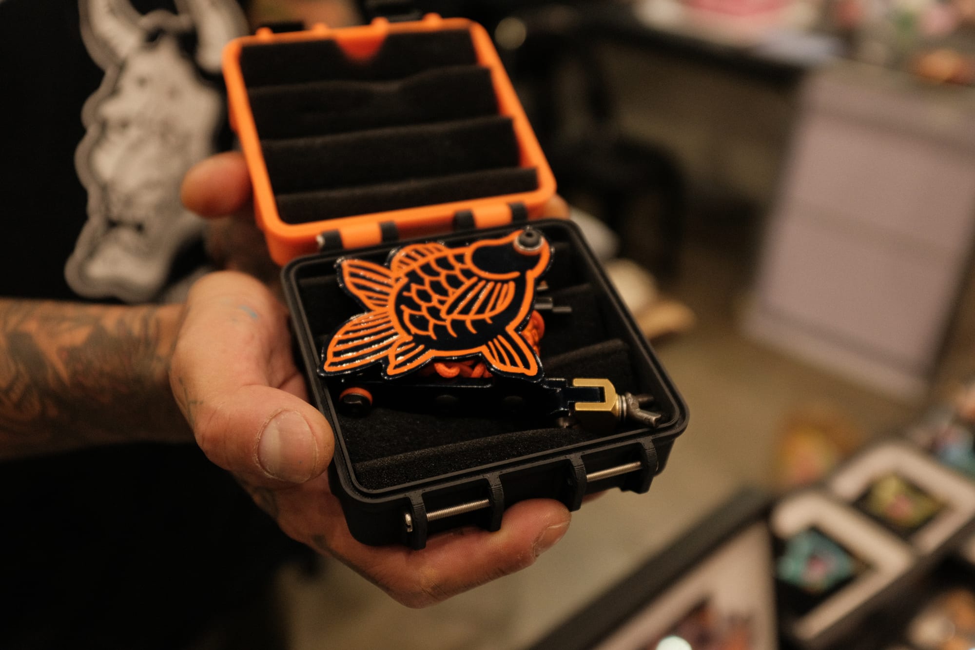 A tattooed hand holds a small black-and-orange case with a goldfish logo tattoo machine, opened to reveal tattoo machine inside.