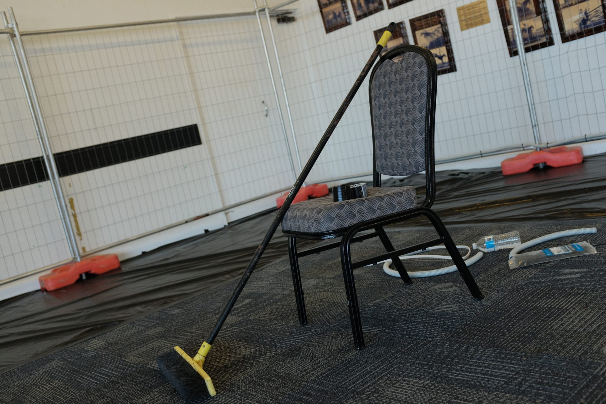 A push-broom leaning on a banquet chair with a roll of black tape on the seat, amid temporary fencing and floor coverings during setup of the Rainforest and Reef expo in Cairns.