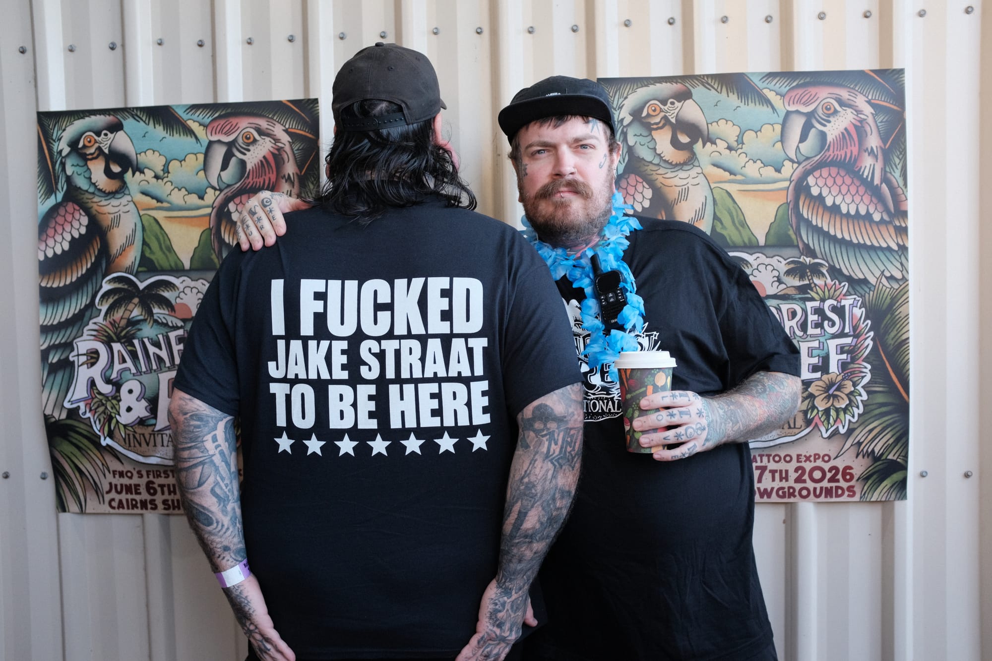 Two tattooed men grin in front of the Rainforest and Reef banner; one wears a T-shirt with a joke slogan about his reason for attending the Cairns tattoo expo.