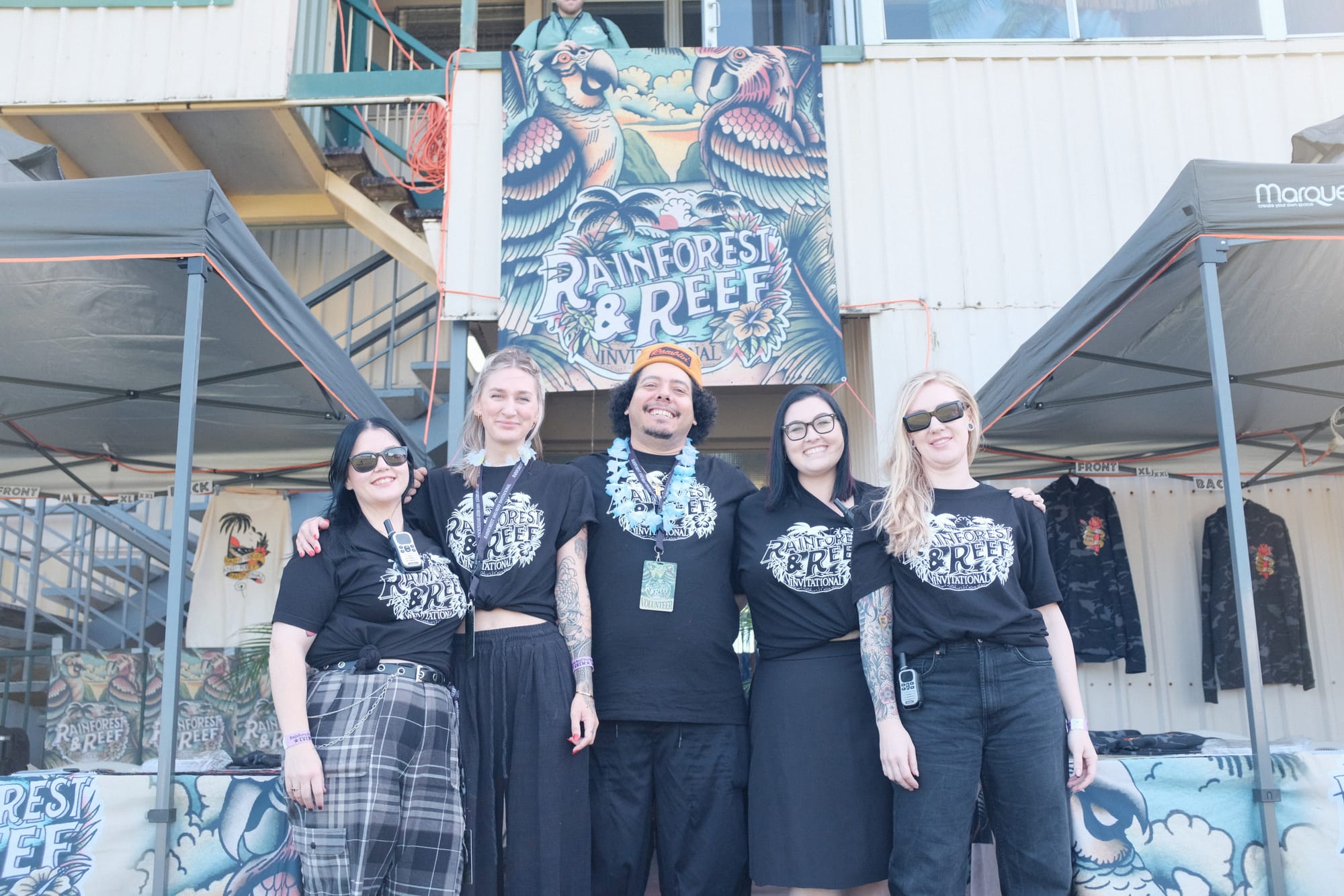 Five Rainforest and Reef event crew members stand smiling in a row in matching black shirts and flower leis beneath the large parrot banner.