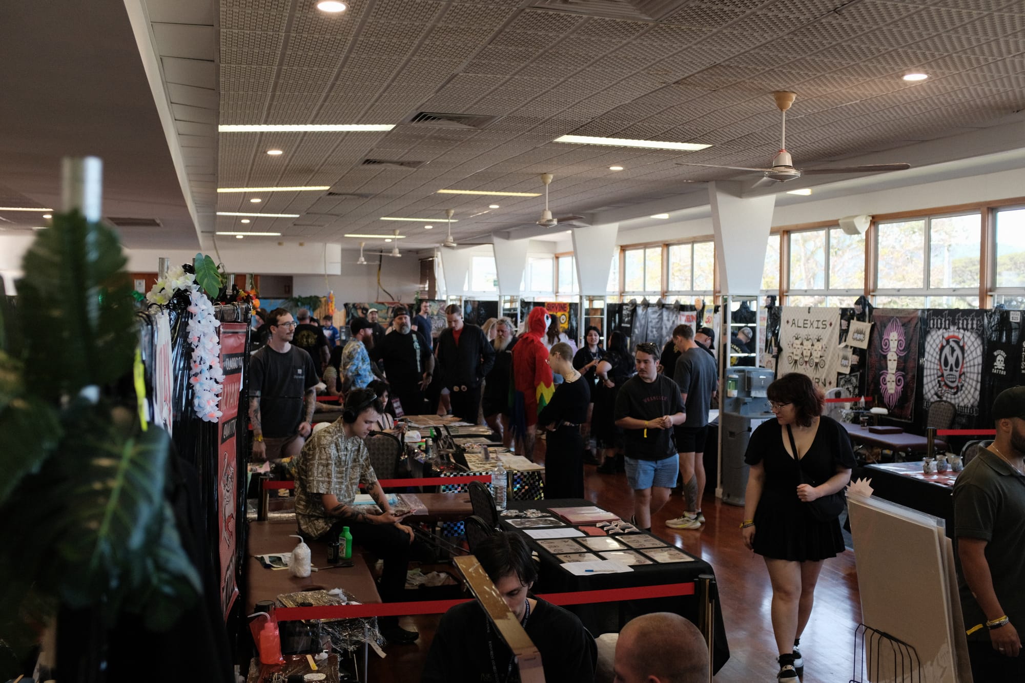 A busy tattoo expo floor on the upper level of the Cairns Showgrounds pavilion, with artists at booths, merchandise banners on the walls and crowds walking the aisles.
