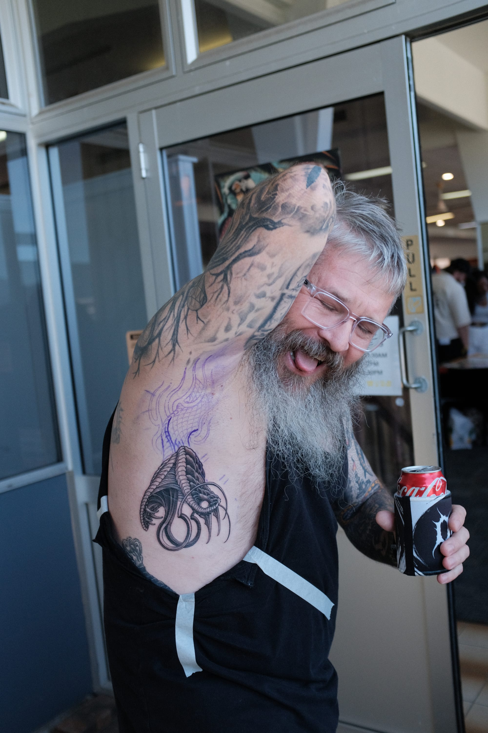 A grinning grey-bearded man in a doorway lifts his shirt to show a fresh black-and-grey cobra tattoo on his ribs, the busy expo floor behind him.