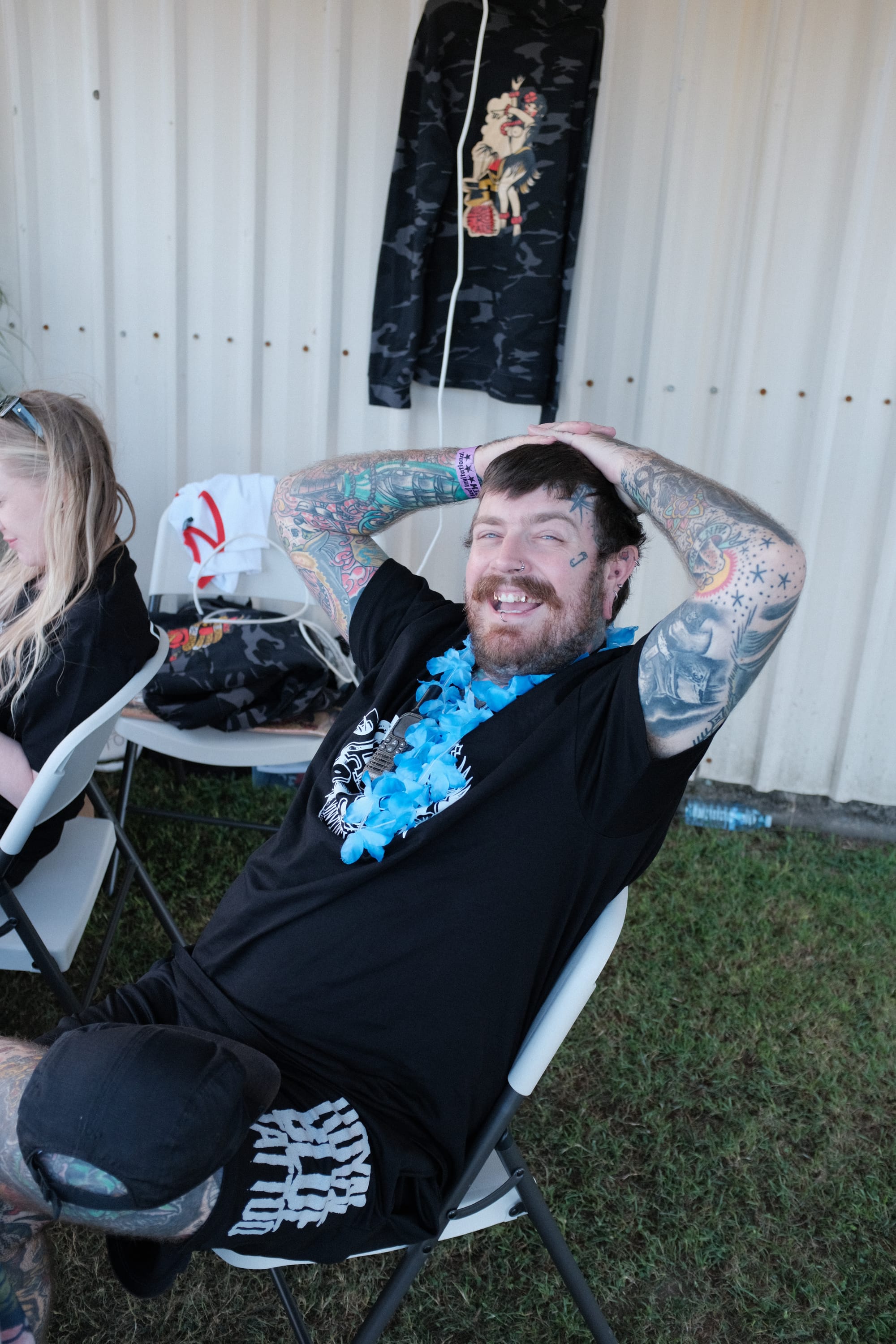 A tired, smiling man in a flower lei leans back in a camp chair with his hands on his head, a colleague seated beside him at the tattoo expo.
