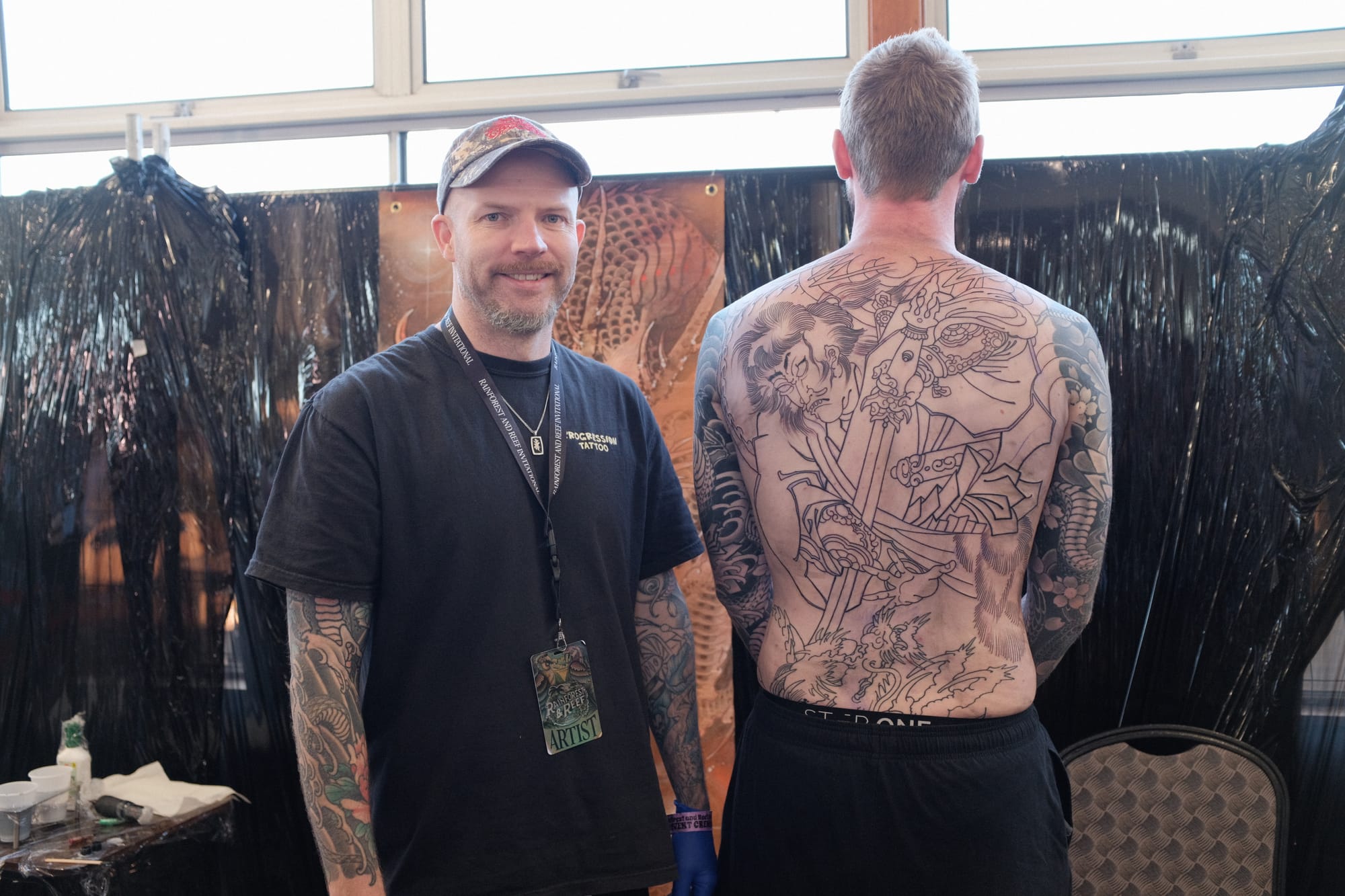 Tattoo artist Sam Hillcoat stands beside a client whose back carries a freshly outlined Japanese bodysuit tattoo at the Rainforest and Reef Invitational in Cairns.