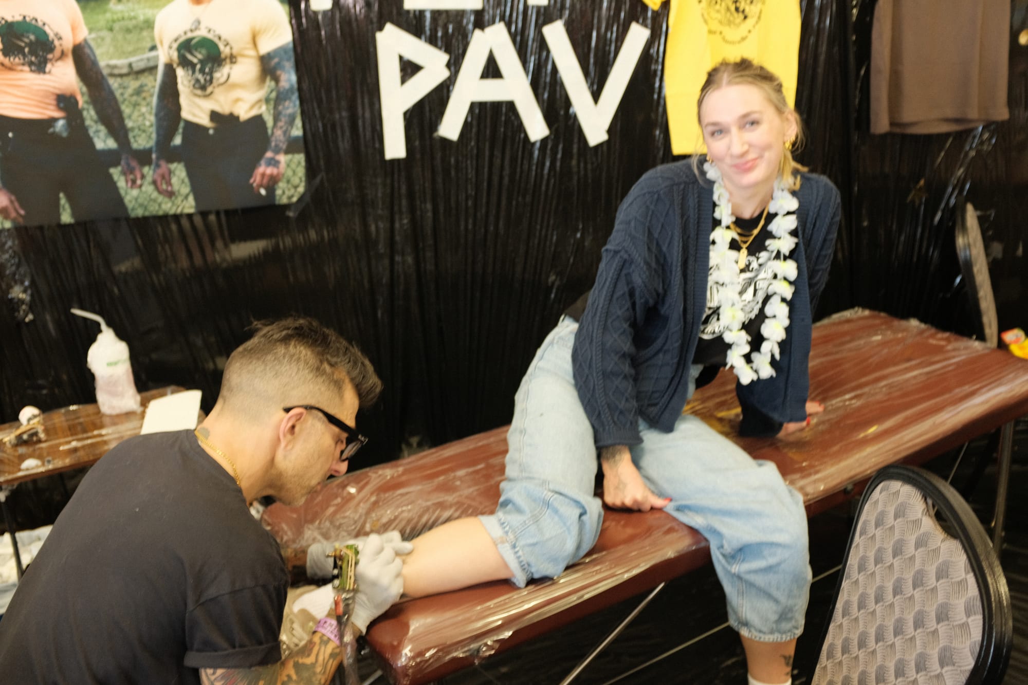 A smiling woman in a flower lei sits on a tattoo table getting a small swallow tattooed on her forearm by artist Pete Pav at his booth during the Rainforest and Reef expo.