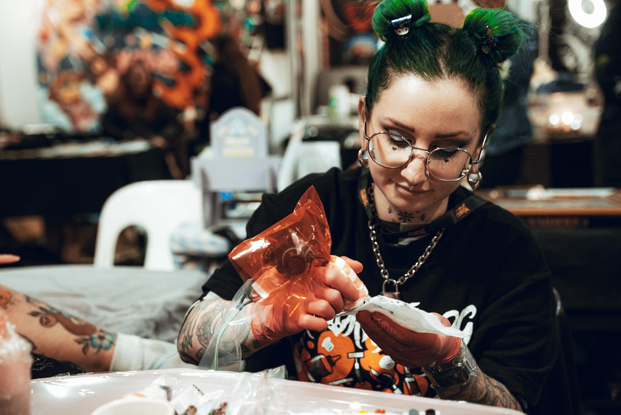 Carly VanVidler, tattooist with green hair in two buns, holding a covered tattoo machine in one hand and a paper towel in the other.