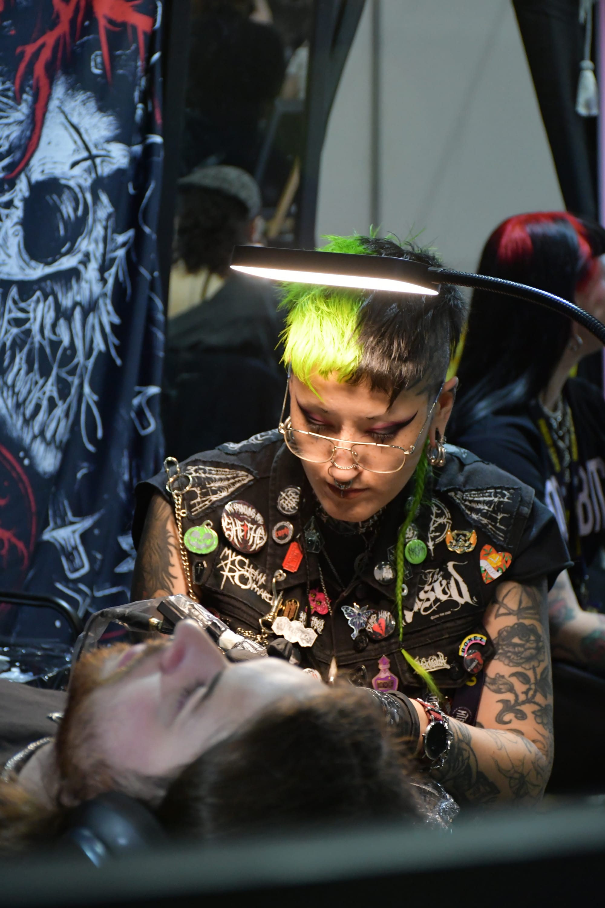 Helen Highwaters, tattooist with green and black hair and a patched vest, tattooing a client under a lamp.