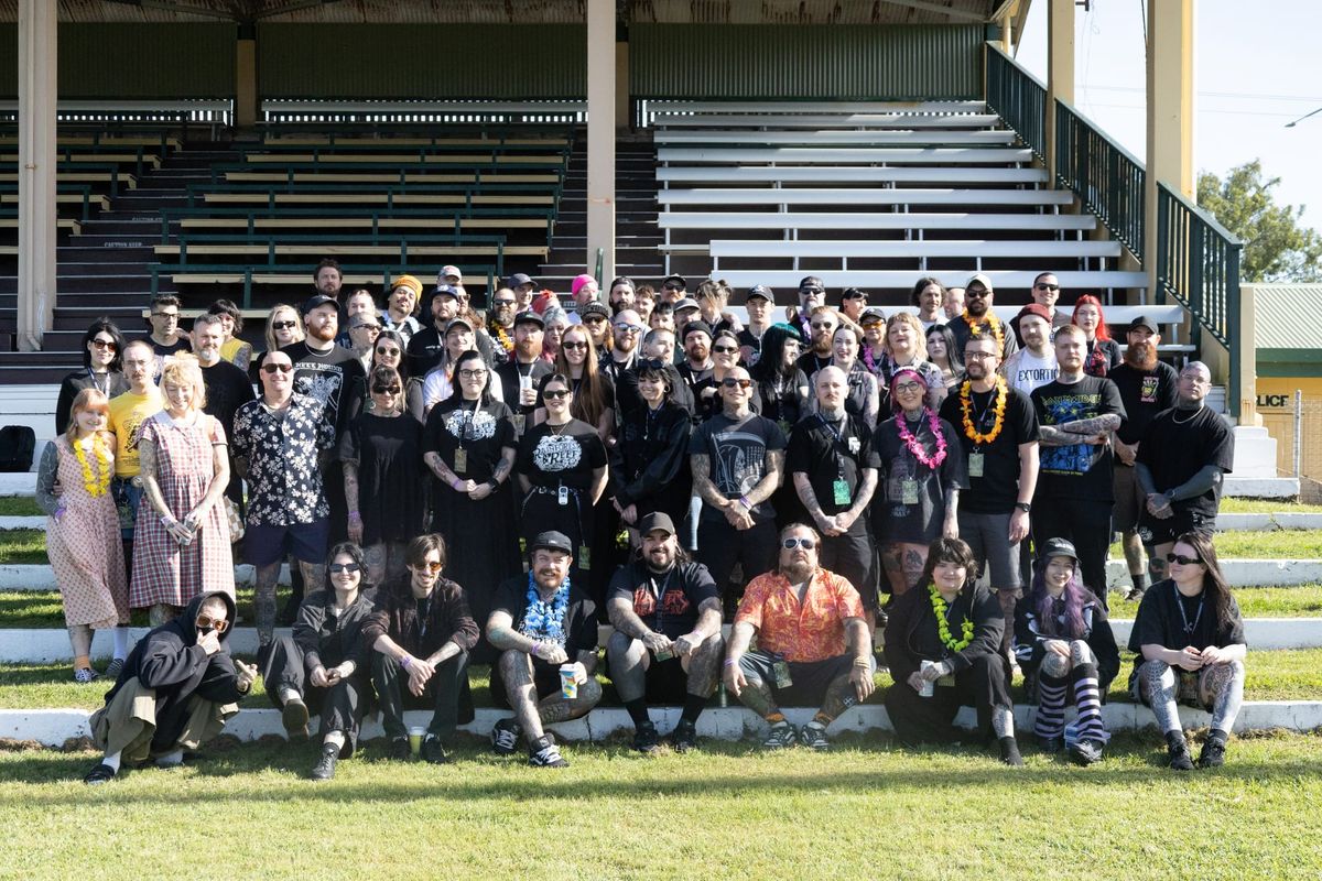 Tattoo artists, vendors and crew pose together on the timber grandstand steps at the Cairns Showgrounds during the Rainforest and Reef Invitational, many wearing leis and band shirts.