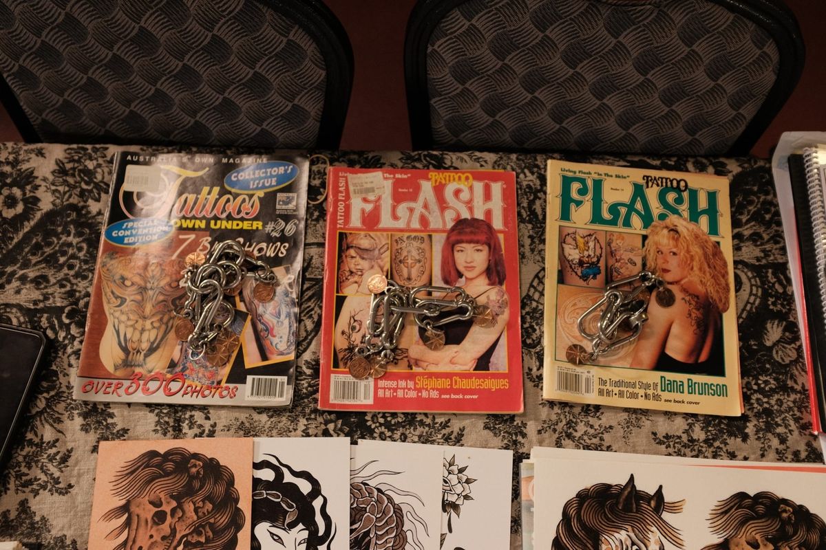 A stack of well-worn vintage tattoo magazines, covers showing tattooed figures, sitting on a cloth-covered table at a tattoo expo.