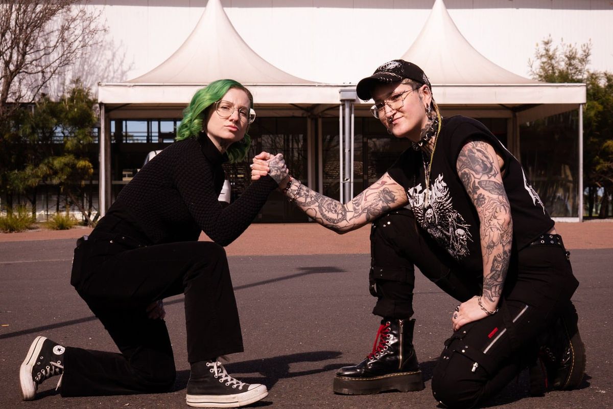 Carly VanVidler, with green hair, and Helen Highwaters, in a black cap, kneeling and clasping hands in front of a white tent-roofed building.