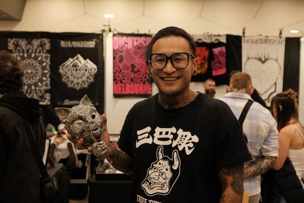 Smiling tattooed man in glasses holds a ceramic cat-head sculpture at his tattoo convention booth, Dickfish Melbourne banners hanging on the wall behind him.