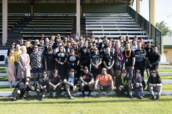 Tattoo artists, vendors and crew pose together on the timber grandstand steps at the Cairns Showgrounds during the Rainforest and Reef Invitational, many wearing leis and band shirts.