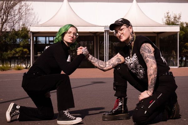 Carly VanVidler, with green hair, and Helen Highwaters, in a black cap, kneeling and clasping hands in front of a white tent-roofed building.