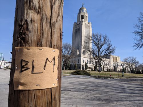the letters BLM embroidered on felt stapled to a utility pole, with the Nebraska State Capitol in the background