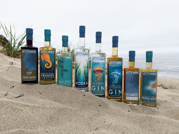 8 bottles of Jettywave liqueurs, gin, vodka, and whiskey in front of a sandy California coastal beach scene
