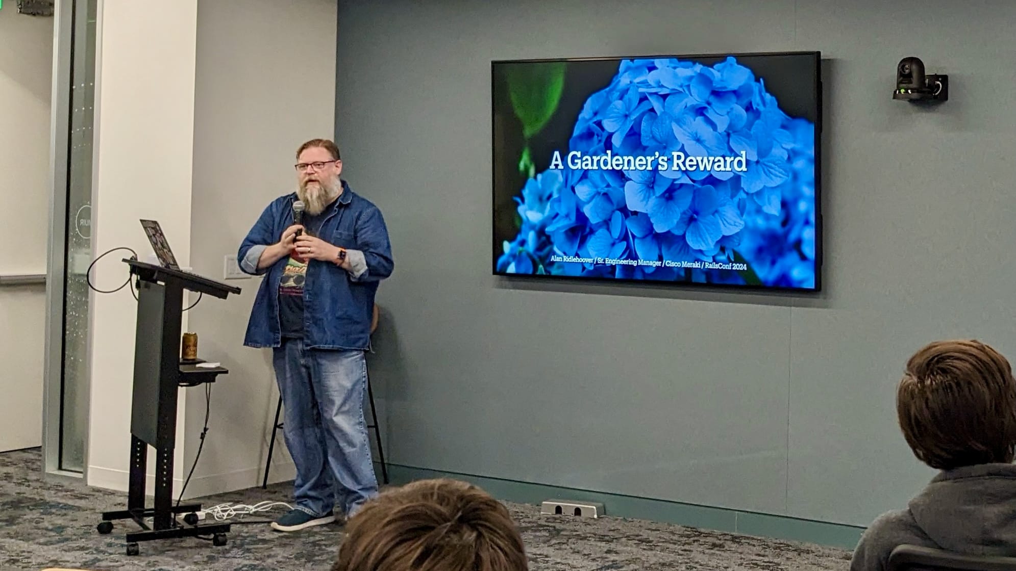 Man in blue denim shirt and jeans talking in front of a television screen with a blue hydrangea and the words "A Gardener's Reward"