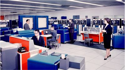 Brightly colored mainframe computer equipment with men and women in suits operating the equipment.