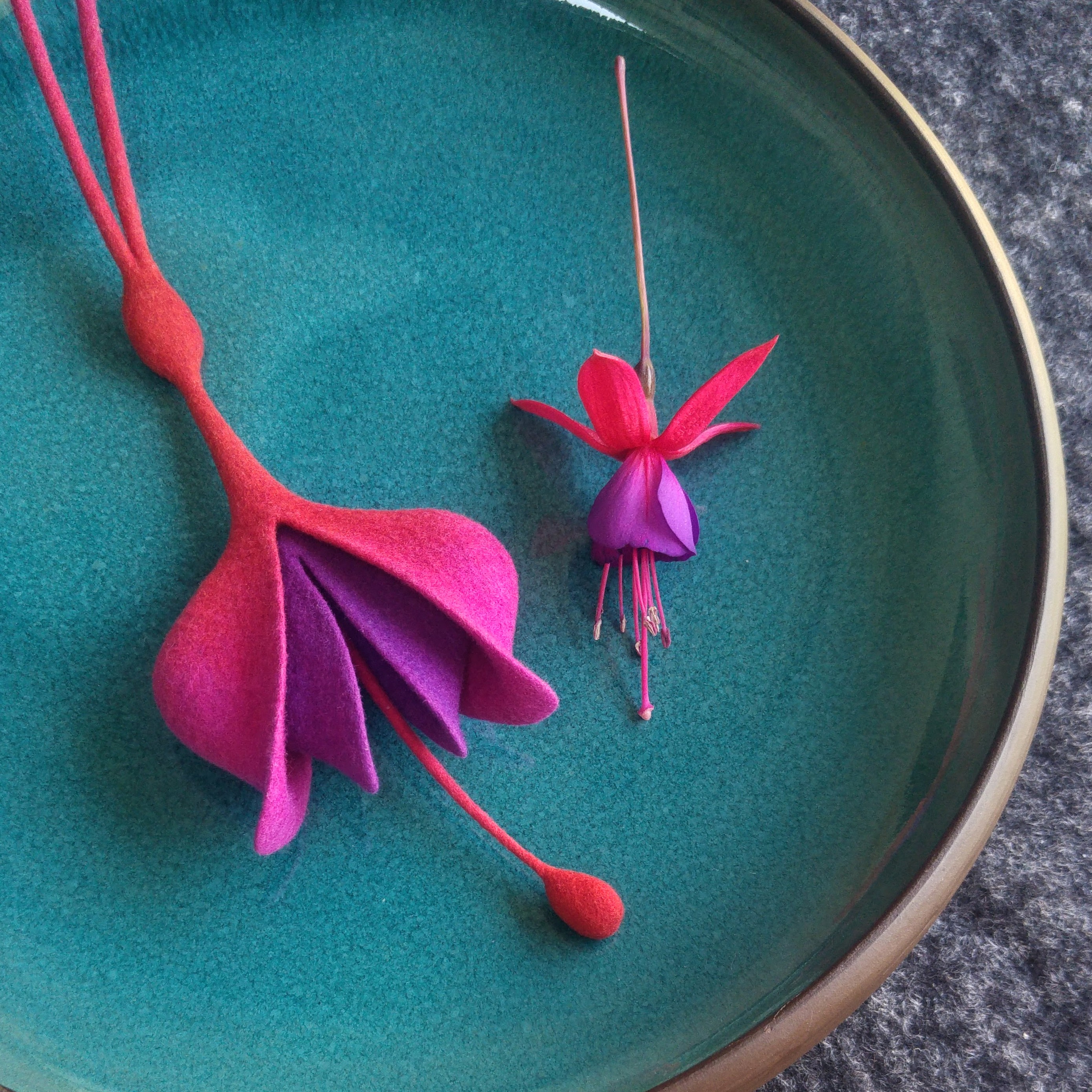 Fuchsia necklace in hot pink and lilac, sitting on a turqouise plate. On the right a real fuchsia flower is sitting for comparison.