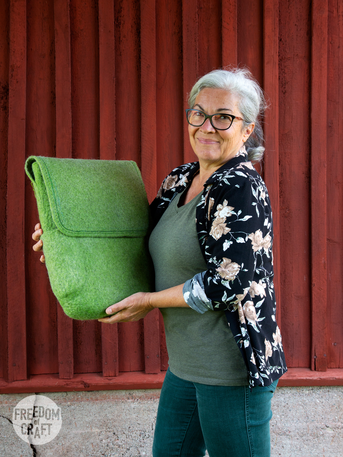 A woman holding her felted green back pack. It is finished. She holds it with pride, and is smiling with satisfaction.