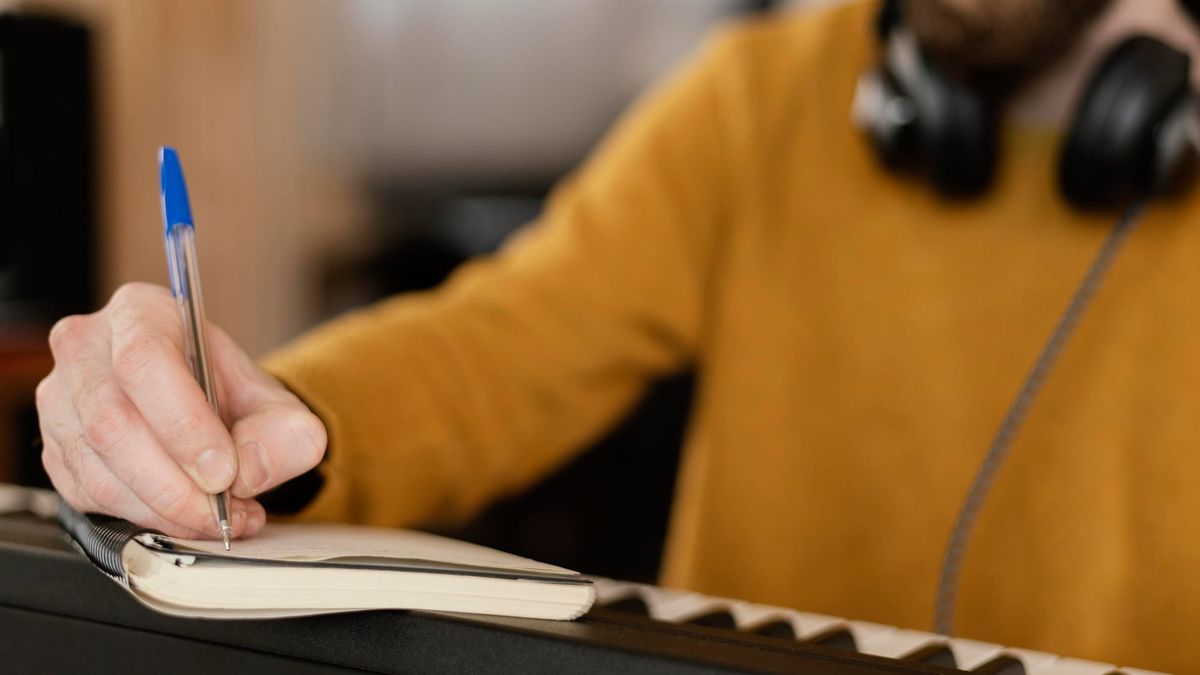 Person writing notes in a notebook while sitting at a keyboard, wearing headphones and a mustard yellow sweater.