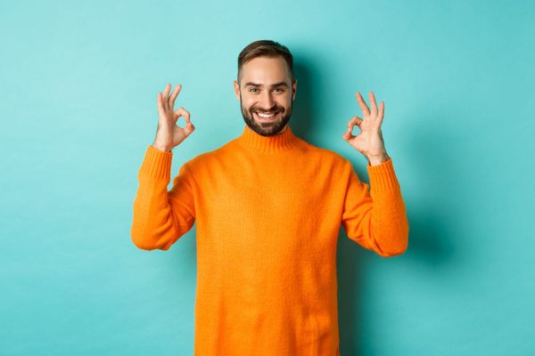 Uomo sorridente con maglione arancione che fa il gesto dell'OK con entrambe le mani su sfondo turchese, esprimendo fiducia e positività