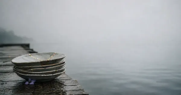 Piatti impilati con bordo riparato in oro, su un molo sull'acqua calma, nella nebbia che si dirada, riflessioni sul dovere