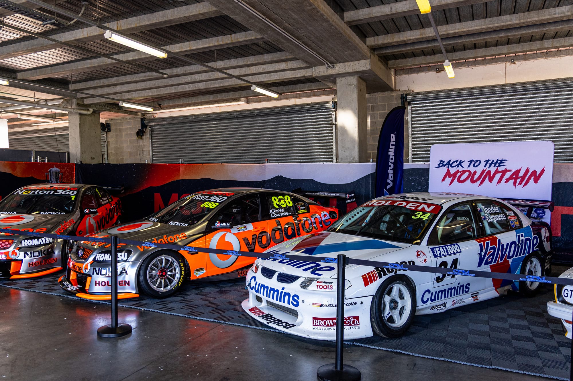Three retired Holden Commodore race cars on display in a pit garage - two in orange and silver Vodafone livery and one in white Valvoline livery - behind a rope barrier with a Back to the Mountain sign.