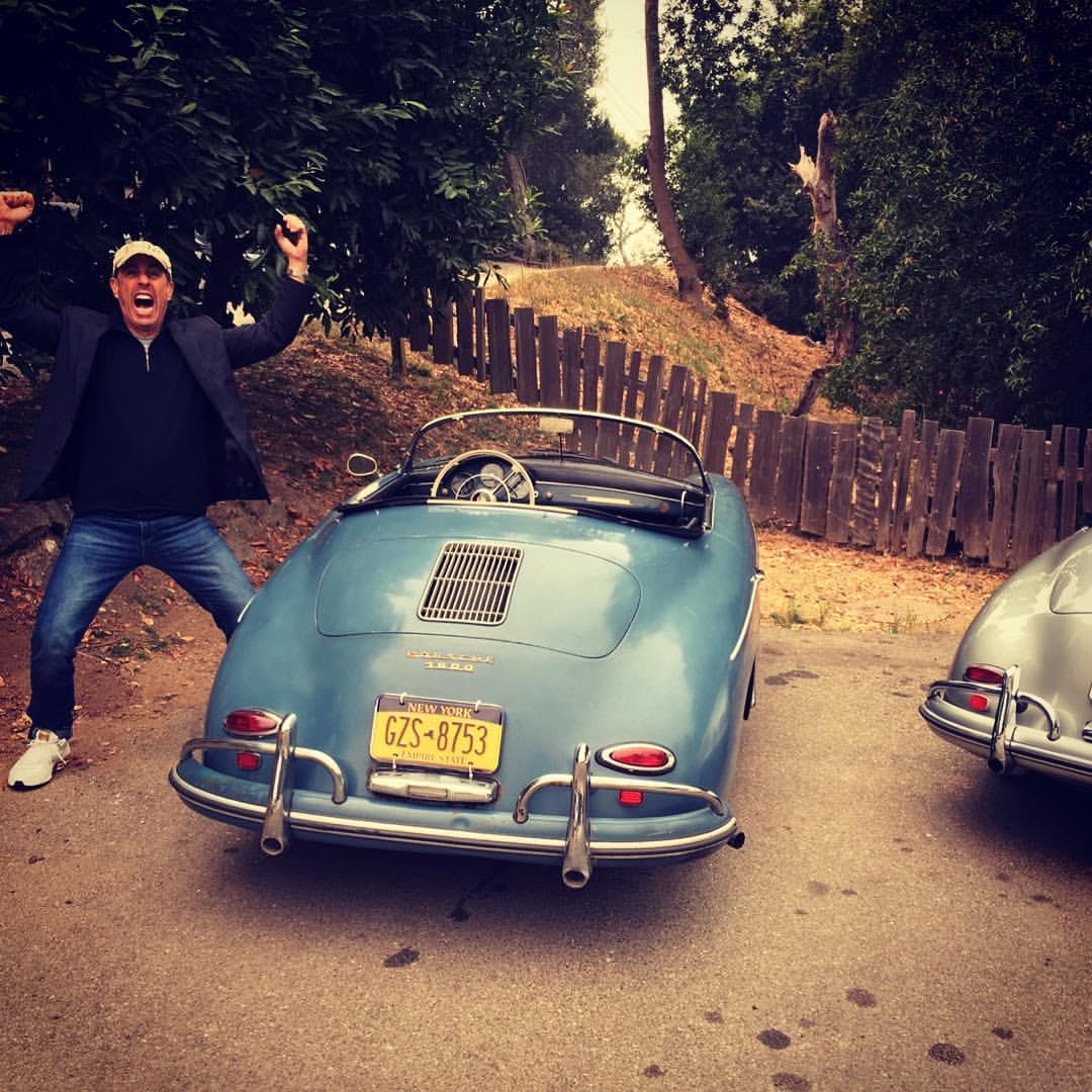 Jerry Seinfeld celebrating beside a light blue 1958 Porsche 356A Speedster with New York plates