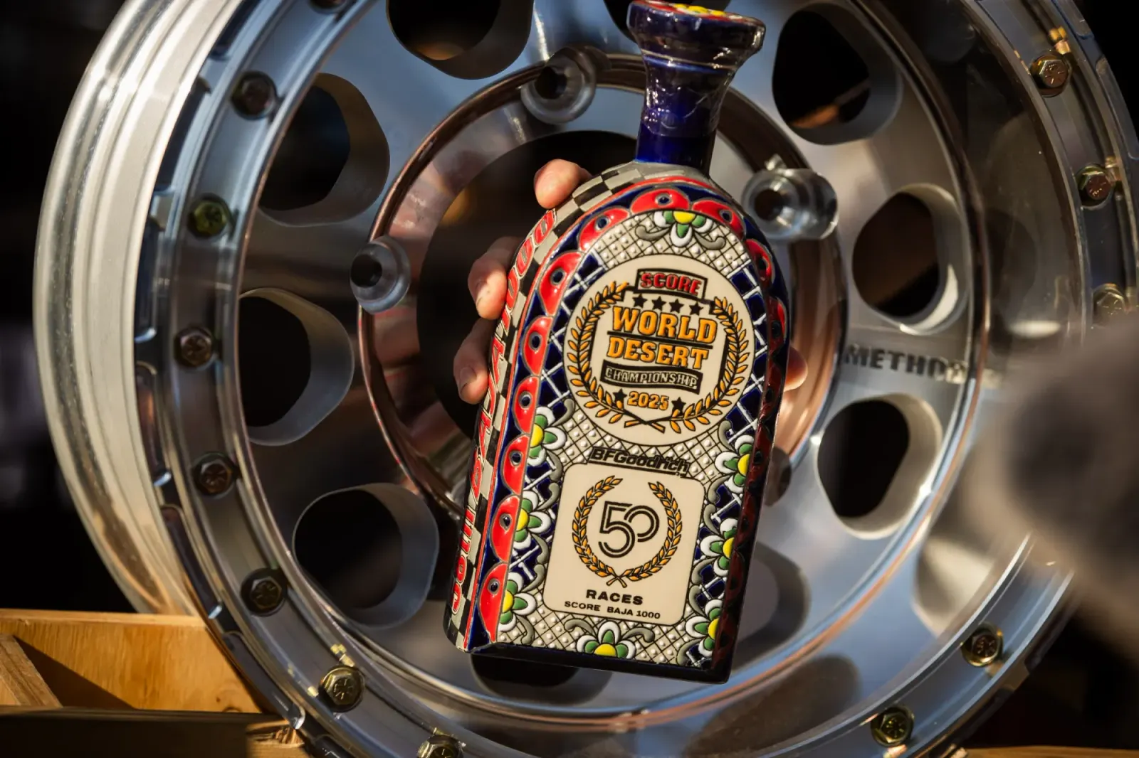 Close-up of a hand placing a colourful ceramic SCORE World Desert Championship trophy in the centre of a polished Method Race Wheels beadlock wheel.