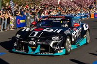 Black and teal number 1 Toyota GR Supra Supercar driven past a large crowd at a street parade, with pink blossom trees behind.