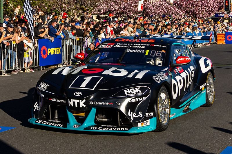 Black and teal number 1 Toyota GR Supra Supercar driven past a large crowd at a street parade, with pink blossom trees behind.