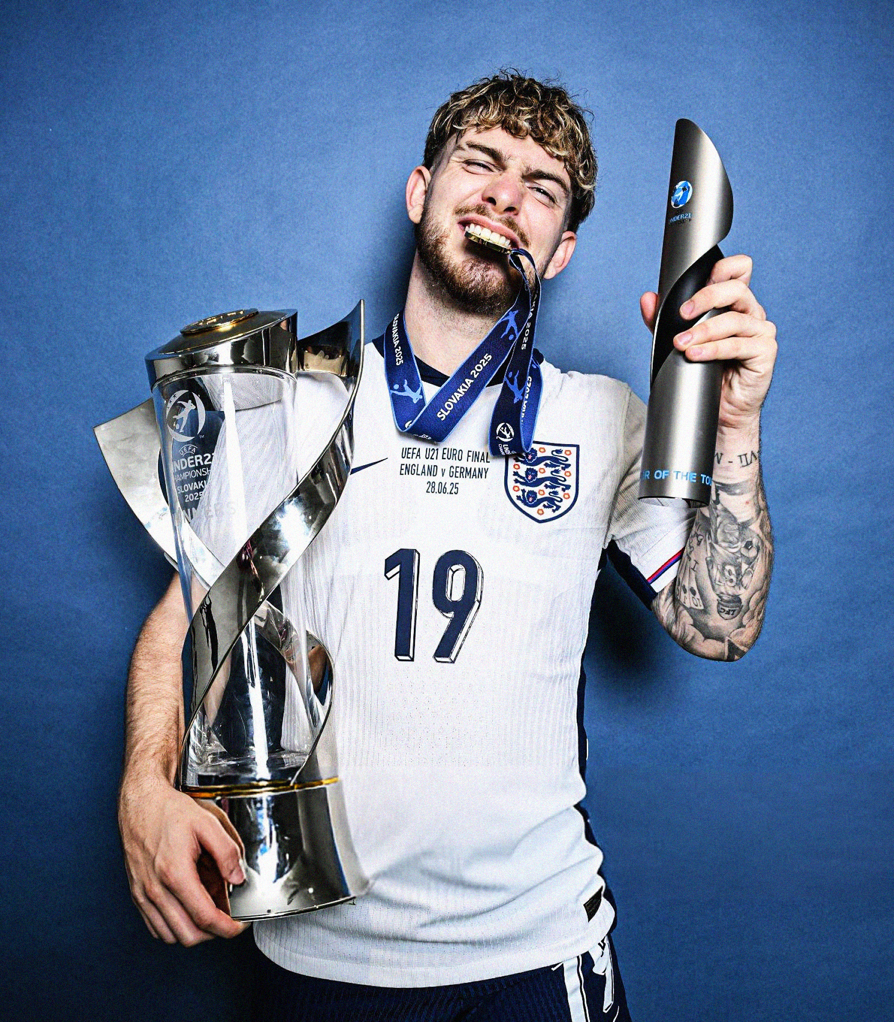 Portrait photo of Harvey Elliott holding the UEFA U-21 EURO and Player of the Tournament trophies while biting his winners' medal, set against a deep blue background