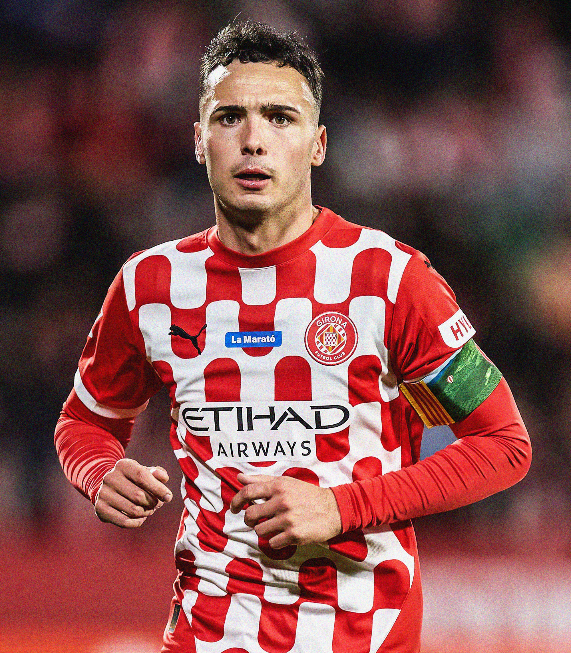 Portrait photo of Arnau Martínez wearing a checkered red-and-white Girona shirt, looking directly at the camera while running toward it