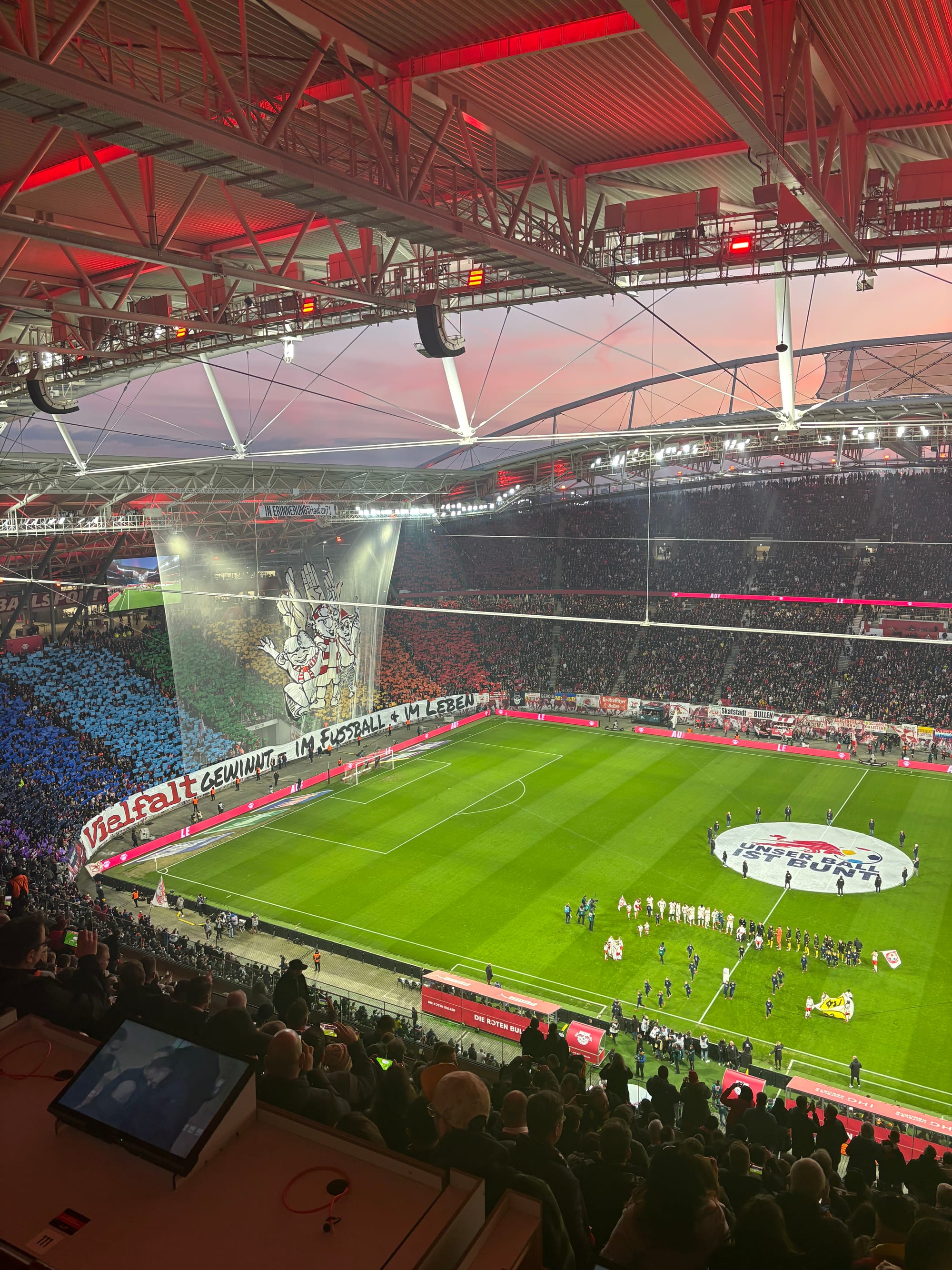 The crowd before kick-off, RB Leipzig vs Borussia Dortmund, from high in the stands - to our left, an immense tifo unfurls, reading in German: 'Diversity wins, in football and in life'. The Leipzig fans behind have formed a pride flag with placards.