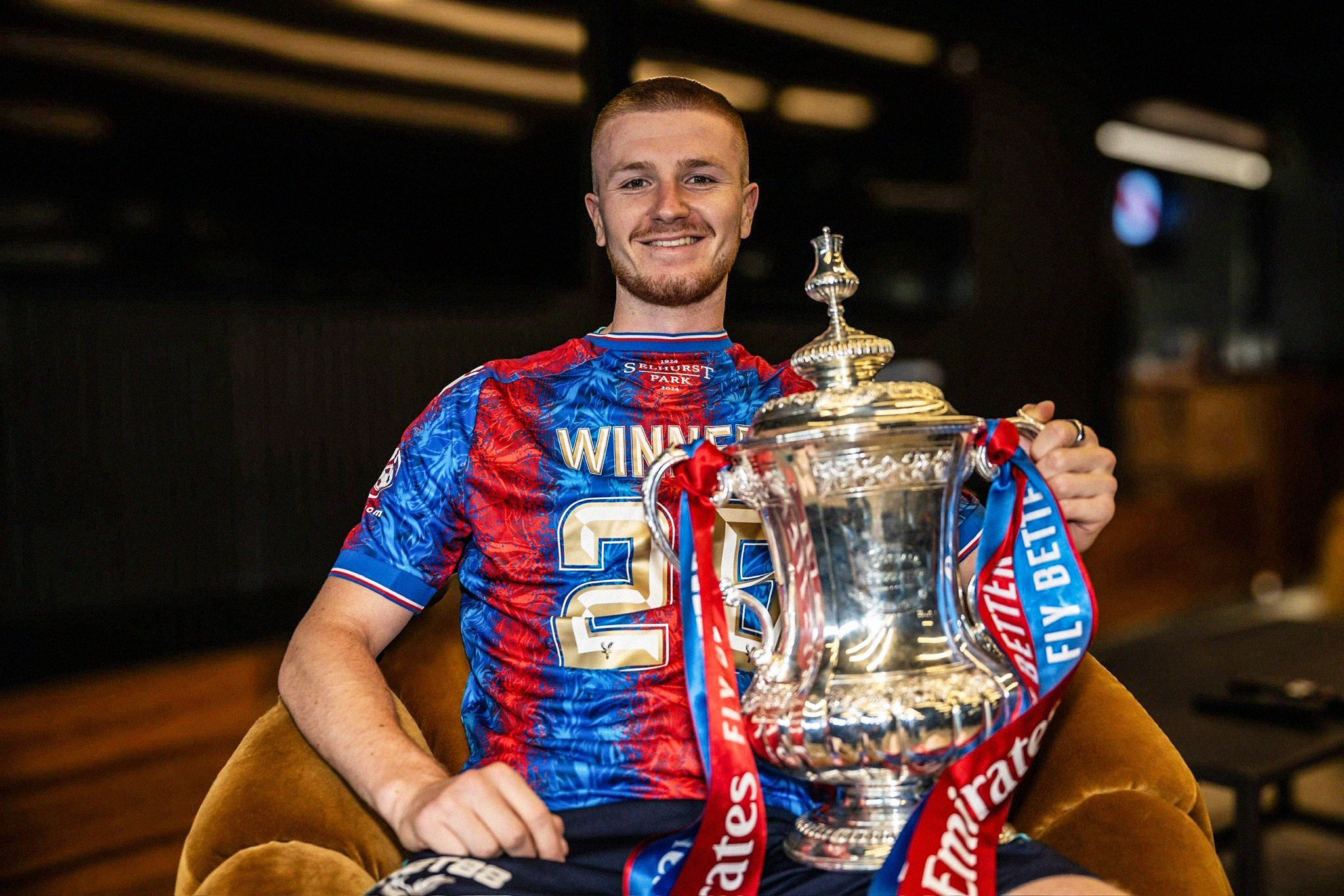 Photo of Crystal Palace's Adam Wharton sat down, smiling at the camera, with the FA Cup trophy on his lap