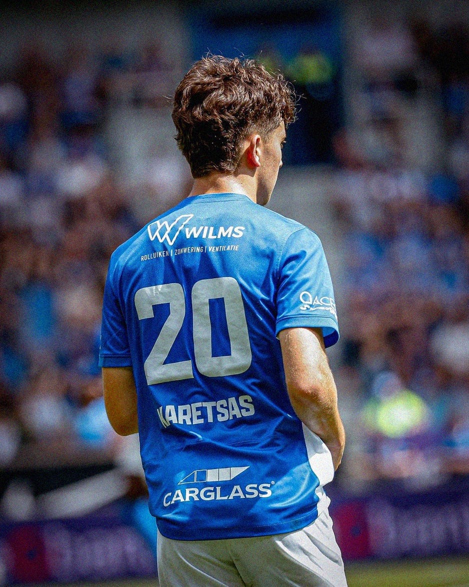 A photo of Konstantinos Karetsas playing for KRC Genk. He's wearing a blue shirt with white shorts. His back is visible to the camera, with 'Karetsas 20' on the back of his shirt.