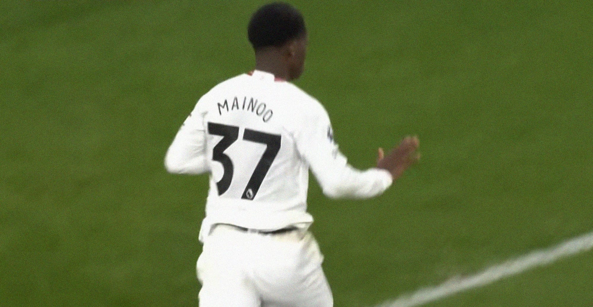 A screenshot of Kobbie Maino celebrating a goal while holding his hand out in a 'calm down' gesture, He's wearing an all-white Manchester United kit with a black typeface on his back, which reads 'Mainoo 37'.
