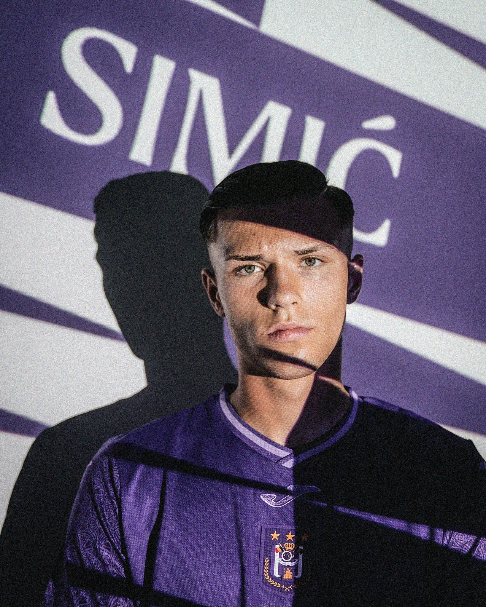 A photo of Jan-Carlo Simić looking directly at the camera, wearing an all-purple Anderlecht shirt. There are different shades of light across his face and upper-body, some bright and some shadow. He's stood in front of a wall with "Simić" projected onto it.
