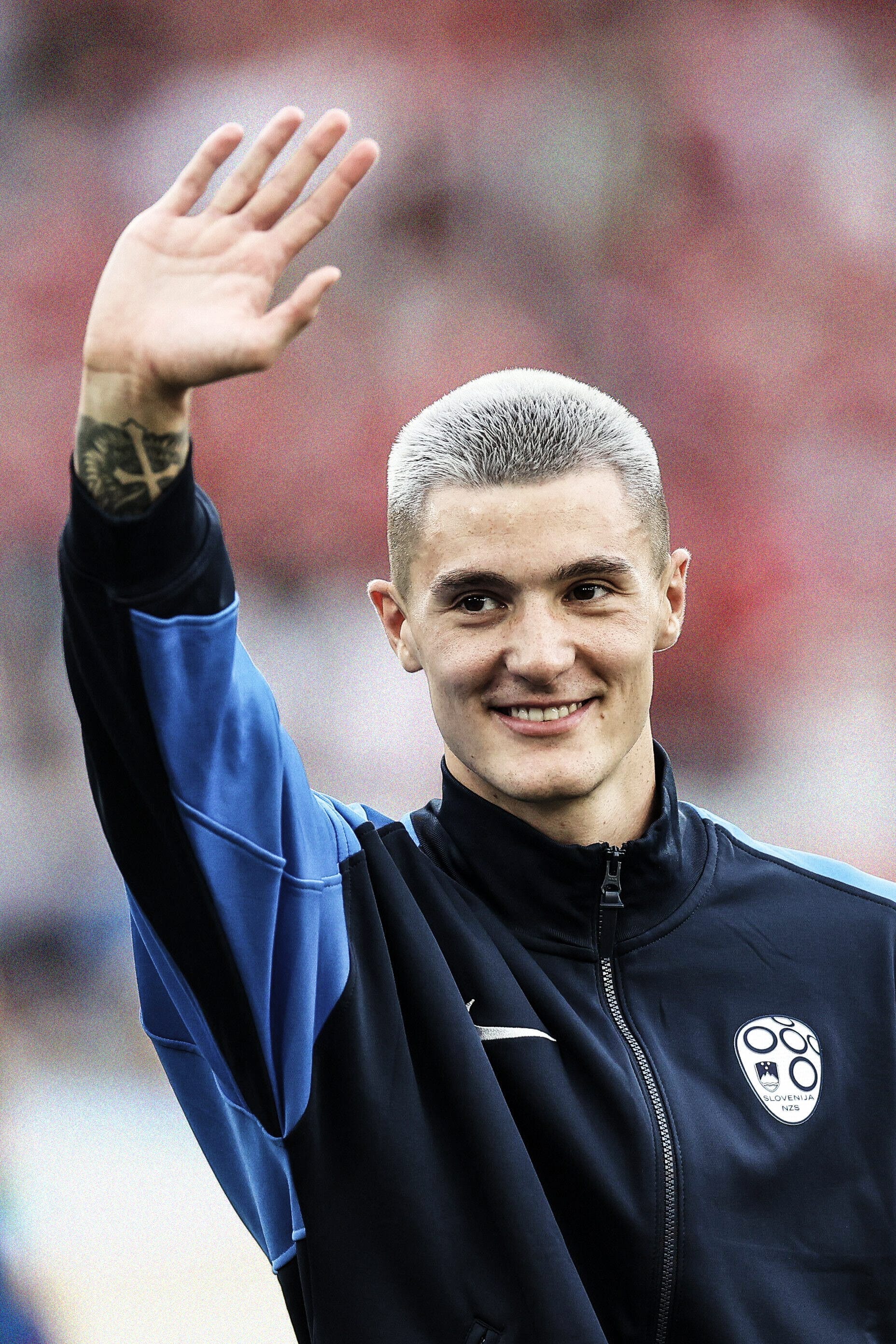 A portrait photo of Benjamin Šeško smiling and waving at EURO 2024, wearing a navy blue Slovenia pre-match jacket