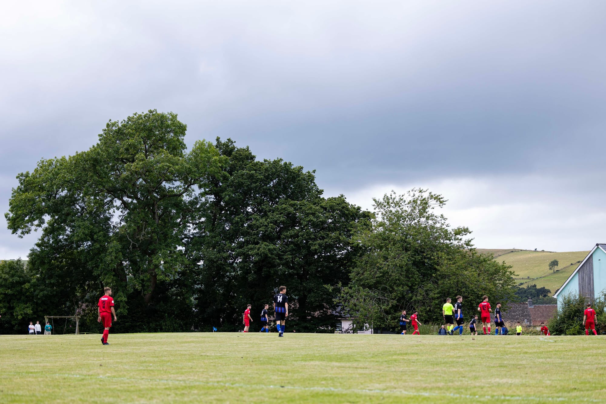 A photo of CPD Felinfach playing Chirk AAA in the FAW Welsh Cup. It shows players on a domed pitch, with large oak trees, council houses and rolling hills in the background.